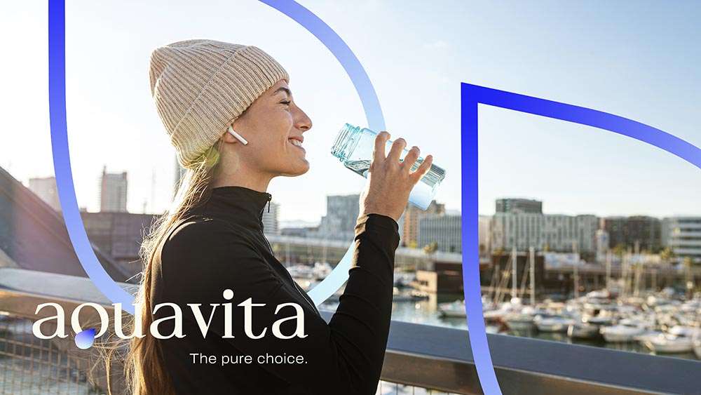 Dia soleado, primer plano de chica sonriente rubia, de pelo largo, con gorra de canalé, se acerca una botella de cristal con agua para beber, al fondo se aprecia un puerto con barcos atracados, tiene un fondo con la silueta vectorial de unas gotas con línea degradado azules y el logotipo 'AQUAVITA. The pure choice' en el lateral izquierdo inferior.