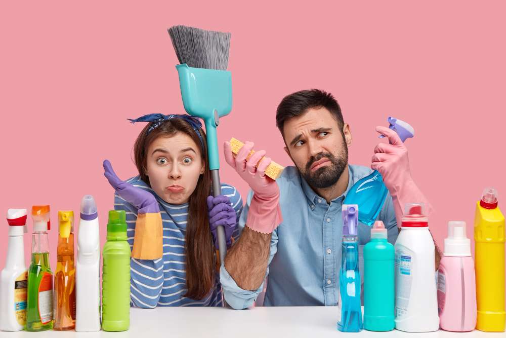 Pareja con guantes de plástico en sus manos rodeada de productos químicos y de utensilios para la limpieza con cara de duda y consternación.