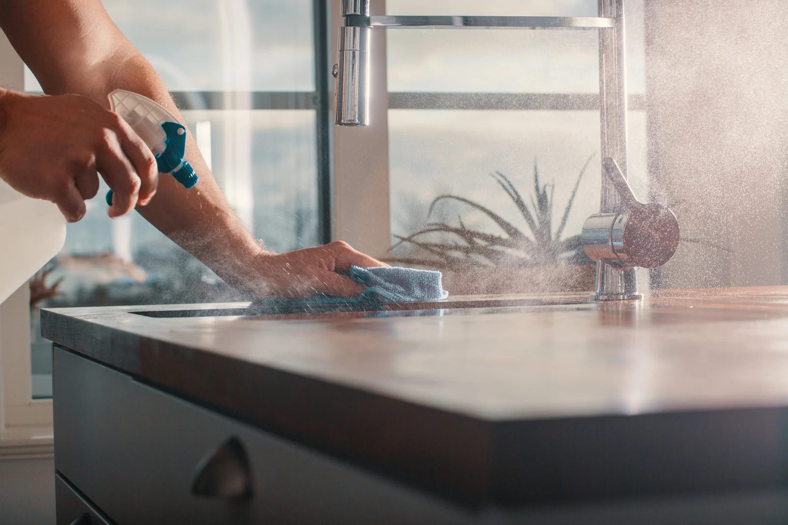 Primer plano de brazos masculinos limpiando una encimera de cocina con un paño de microfibra y un pulverizador.