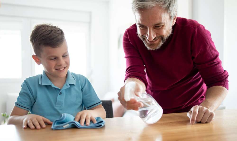 Padre e hijo con caras felices limpiando una mesa con una mopa y un spray que contiene agua ozonizada.