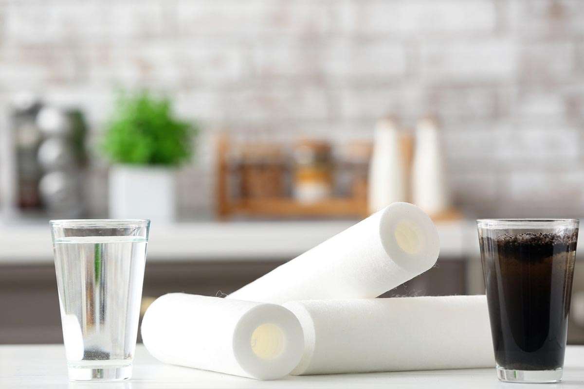 En una cocina, sobre una encimera blanca, hay dos vasos, uno con agua limpia y otro con sucia y entre medio unos filtros de osmosis.