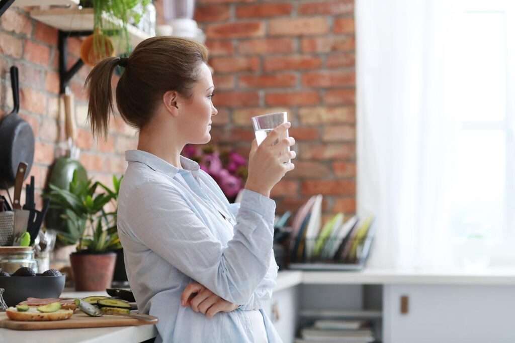 Mujer con apariencia relajada de pie en una cocina bebiendo agua filtrada por osmosis y con comida saludable en una encimera que tiene al lado.