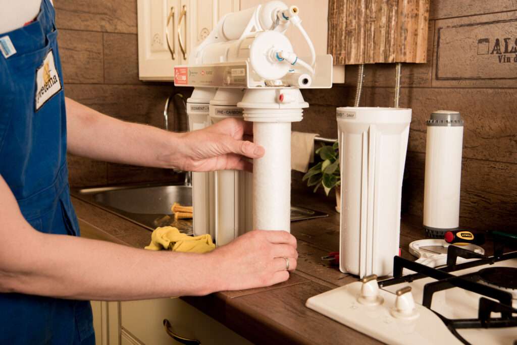Un operario cambia filtros de sistema de agua por osmosis en una cocina clásica donde se ven muebles de madera, fogones de gas y un lavadero con el grifo de salida de agua purificada.