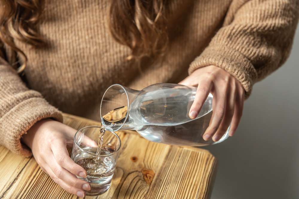 Mujer rellena un vaso con agua de una jarra encima de una encimera de madera.
