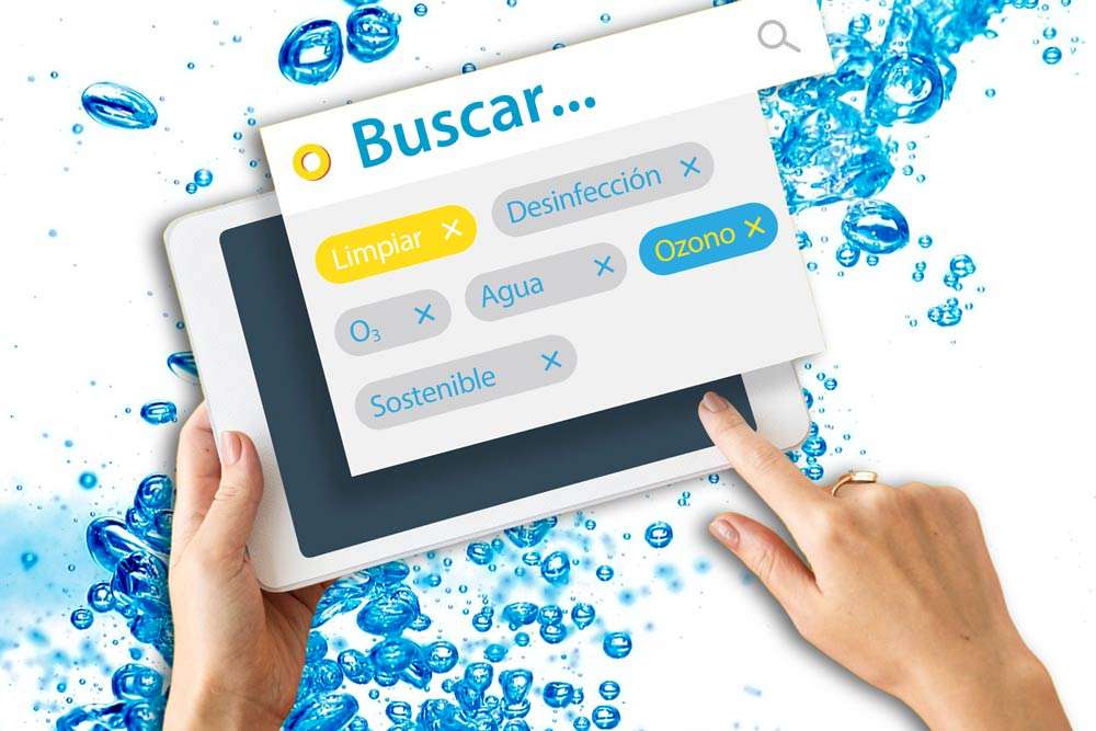 Fotomontaje con fondo de burbujas de agua azules con unas manos en primer plano que sujetan y manipulan una tablet donde sobresale una imagen de búsqueda con palabras clave 'Ozono', 'O3', 'Limpiar', 'Desinfección' 'Agua' y 'Sostenible'. La imagen es de colores claros y muy luminosa