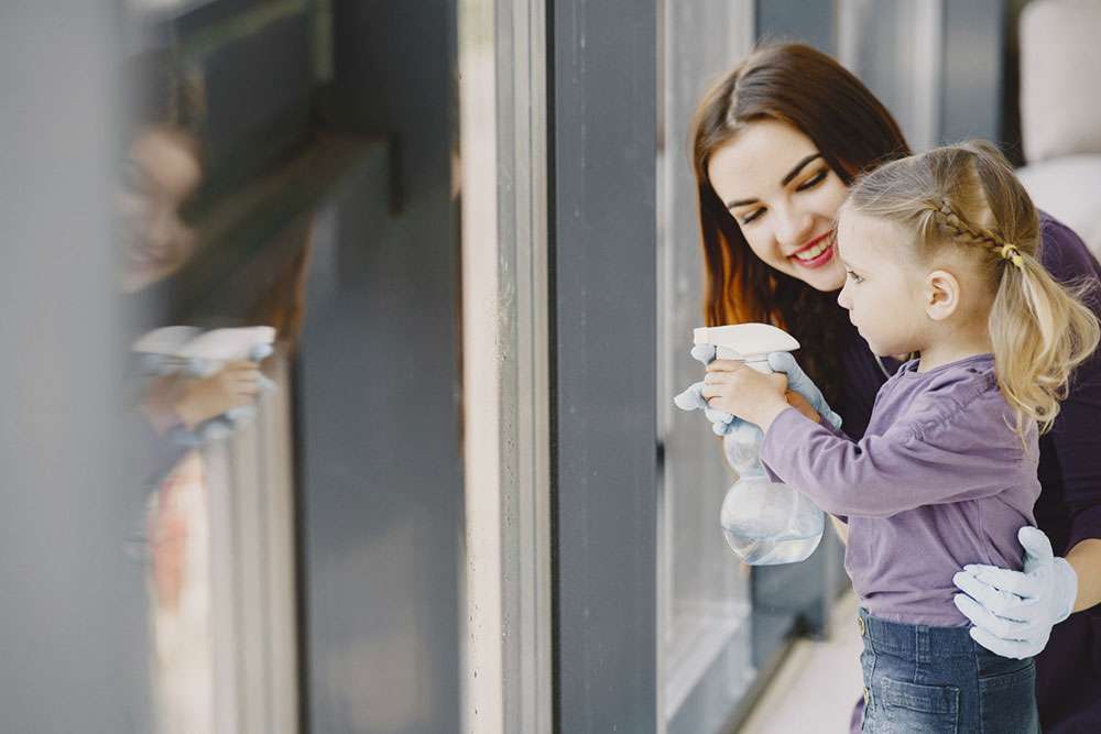 Una madre feliz y una niña limpian unos ventanales con un pulverizador con agua que sujeta la pequeña.