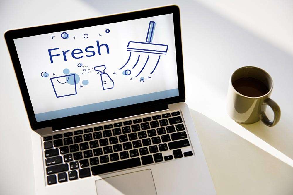 Sobre una mesa hay una taza con café y un ordenador portátil donde se ve en la pantalla la palabra 'fresh' y unas ilustraciones de un cubo de fregar, un pulverizador y una mopa rodeados de burbujas y estrllas que evocan brillo y limpieza.