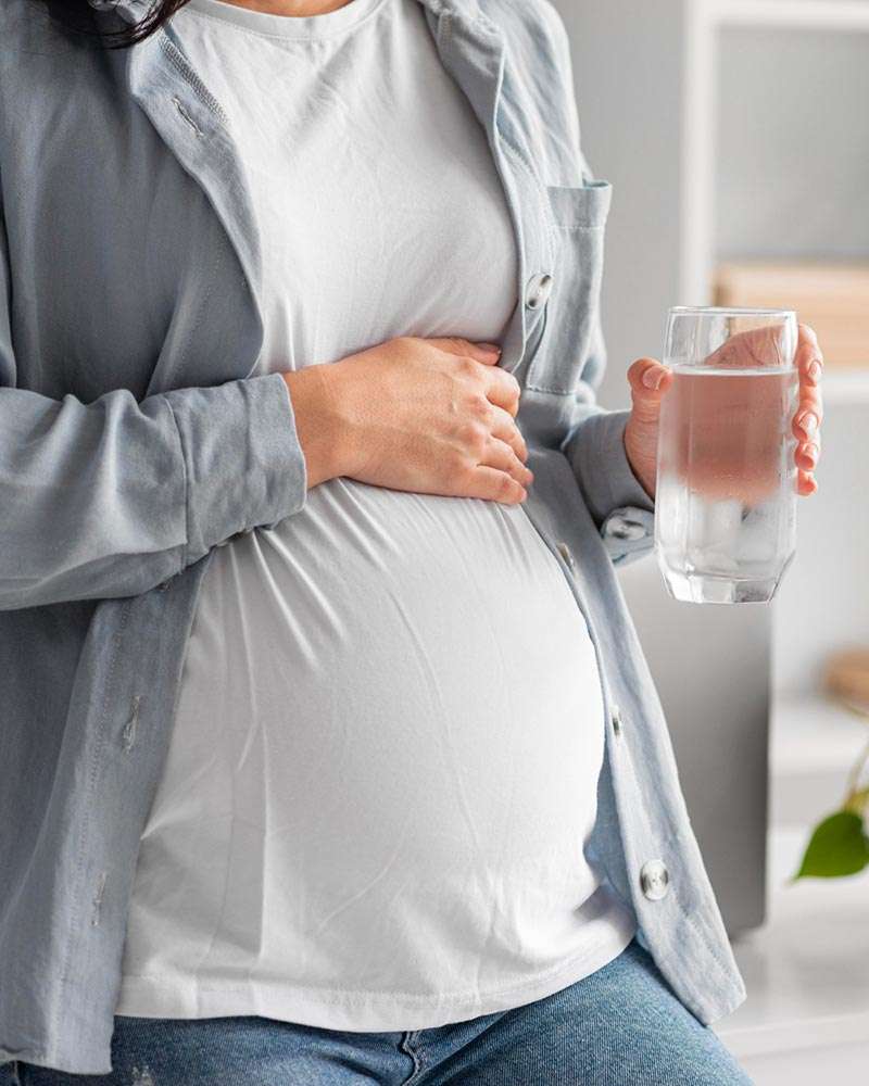 Mujer embarazada tocándose la barriga sujeta vaso largo de agua. Imagen muy luminosa de tonos claros.