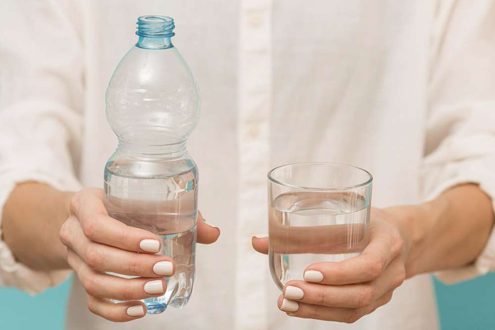 Imagen luminosa de primer plano de manos de mujer sujetando a su izquierda una botella de plástico medio llena de agua y a la derecha un vaso lleno de agua.