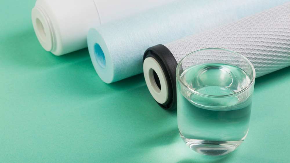 Tres tubos diferentes de filtrado por osmosis junto a un vaso lleno de agua apoyados sobre una superficie de color verde menta
