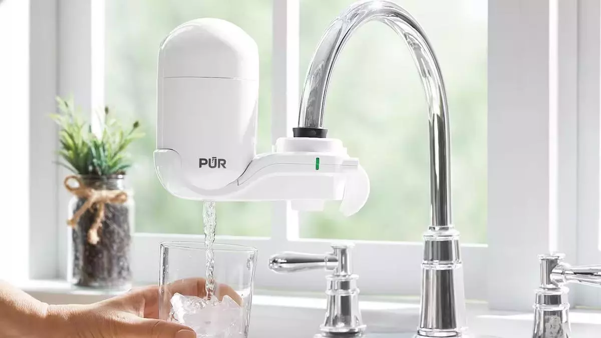 Primer plano salpicadero con ventana luminosa de fondo, una mano sujeta un vaso mientras se llena con agua filtrada con dispositivo PUR osmosis conectado al grifo.