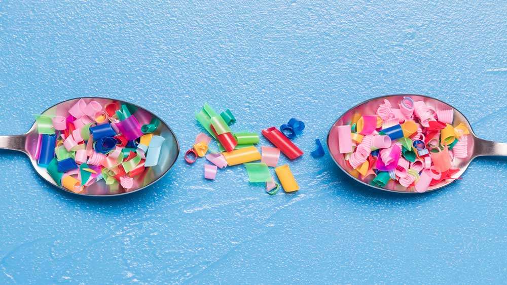Sobre fondo azul, primer plano de dos cucharas planas enfrentadas, con partículas de plásticos de diferentes tamaños, formas y colores en ellas y algunas por fuera de su superficie.