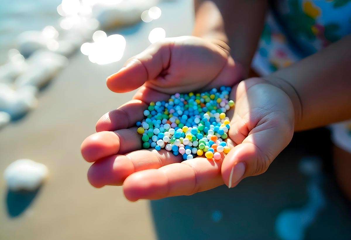 Primer plano de un par de manos sosteniendo gránulos de microplástico de colores con un fondo de playa con arena y agua