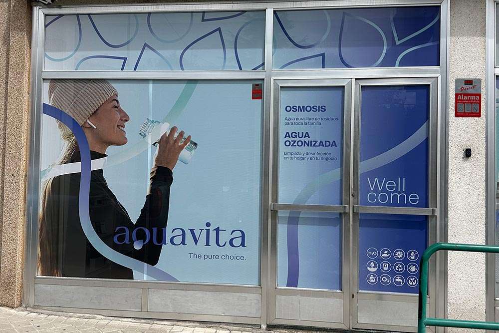 Plano detalle la fachada del local de AQUAVITA con detalle del ventanal con puerta de acceso, mostrando una chica joven deportista bebiendo de una botella.