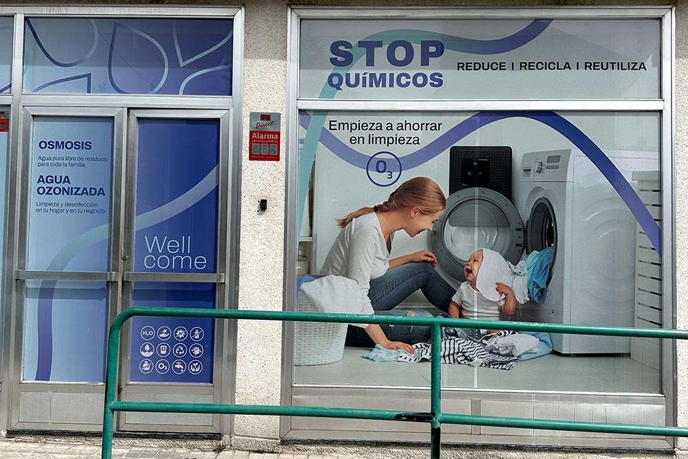 Plano detalle la fachada del local de AQUAVITA con detalle del ventanal mostrando una chica joven sonriente con un bebe sentados en el suelo con una lavadora y una máquina de agua ozonizada ELEKTRA by ECOFROG.