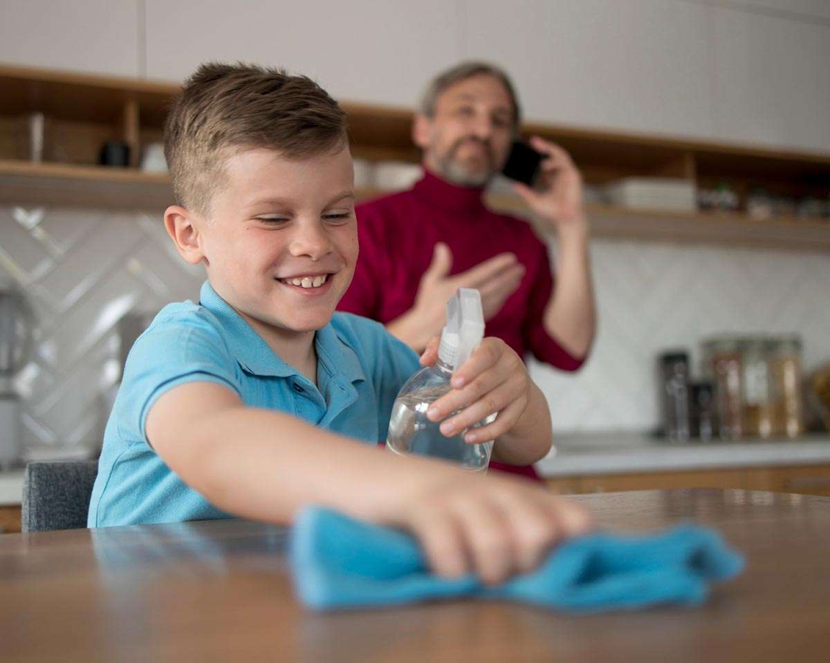En una cocina un padre habla por teléfono móvil mientras su hijo limpia una mesa con una mopa y un spray que contiene agua ozonizada.